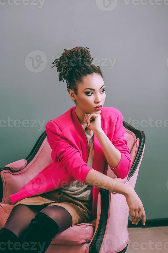 alegre mujer afroamericana en chaqueta rosa sentada en la silla rosa interior. moda, navidad, año nuevo, felicidad, concepto de vacaciones foto