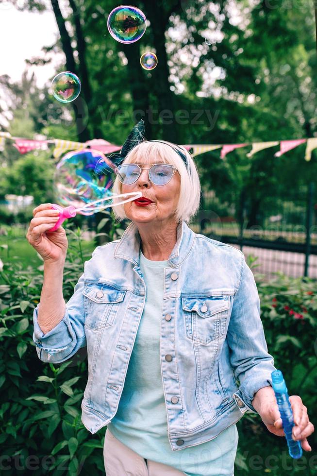 senior stylish woman with gray hair and in blue glasses and jeans jacket blowing bubbles outdoors. Holidays, party, anti age, fun concept photo