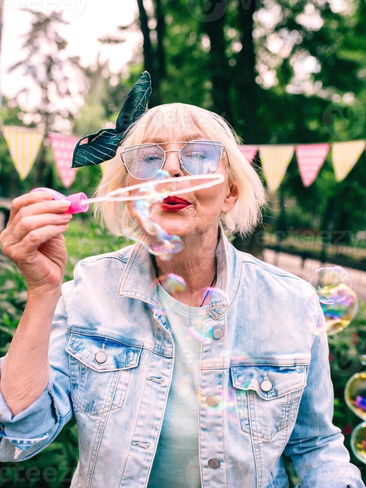 senior stylish woman with gray hair and in blue glasses and jeans jacket blowing bubbles outdoors. Holidays, party, anti age, fun concept photo