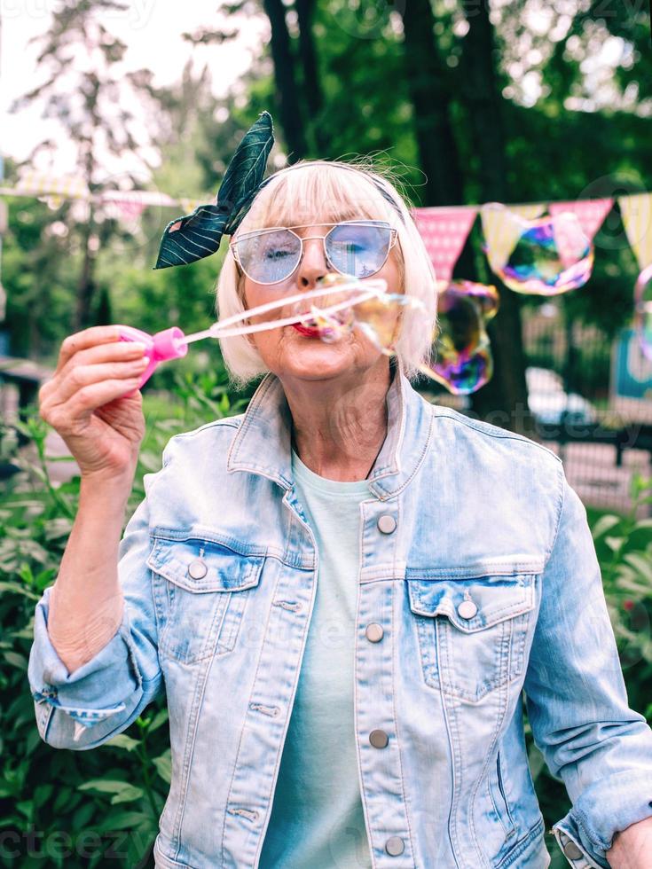 Mujer con estilo senior con cabello gris y gafas azules y chaqueta de jeans soplando burbujas al aire libre. vacaciones, fiesta, anti edad, concepto divertido foto