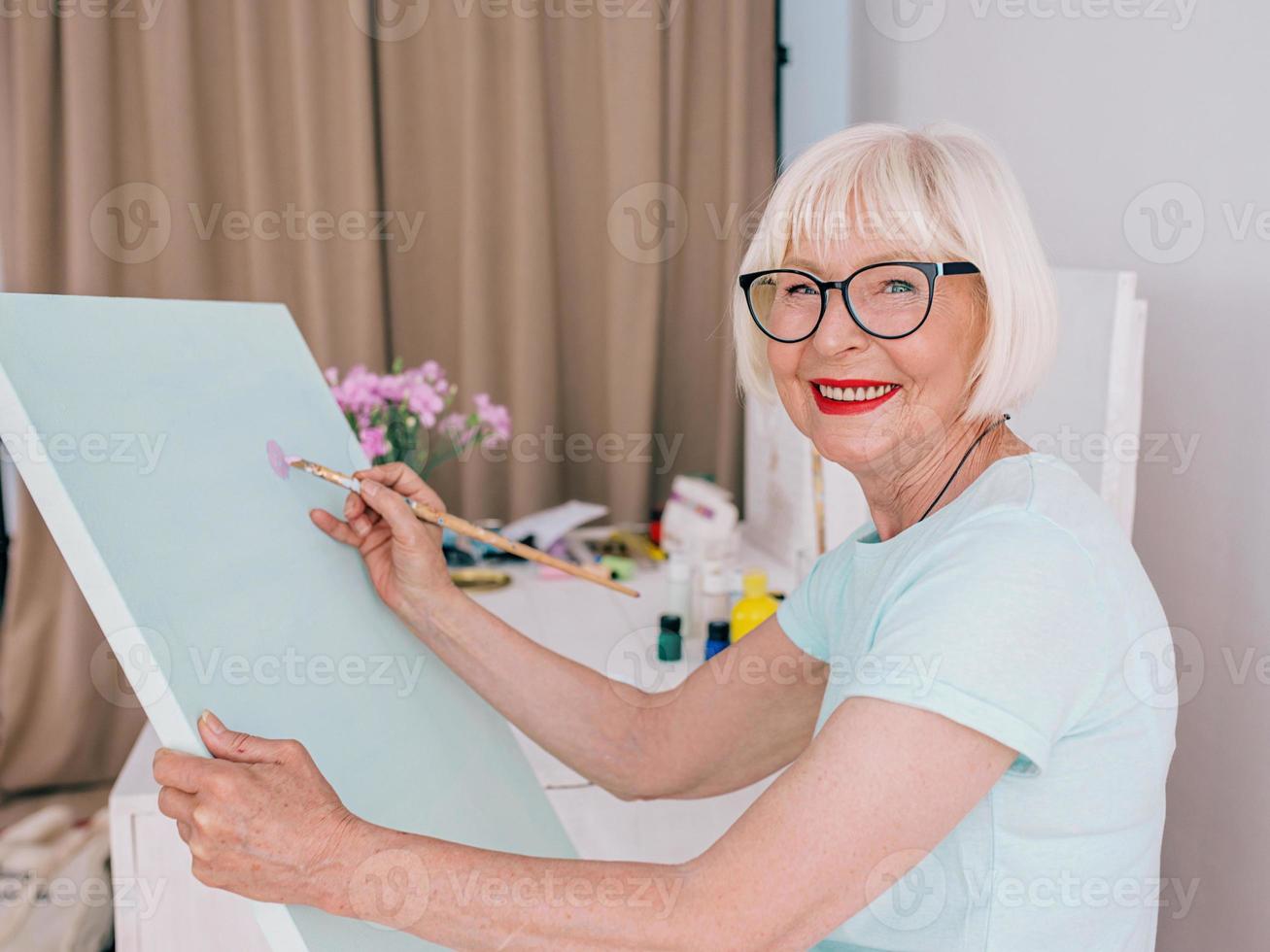 mujer alegre senior en vasos con cabello gris dibujo con lápiz flores en florero. creatividad, arte, pasatiempo, concepto de ocupación foto