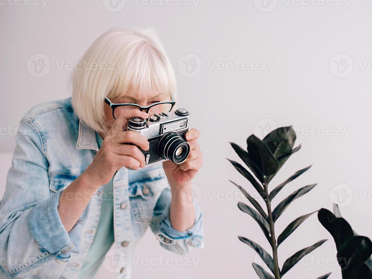 mujer elegante con cabello gris y chaqueta de jeans y gafas tomando fotografías con cámara de película. edad, hobby, anti-edad, vibraciones positivas, concepto de fotografía foto