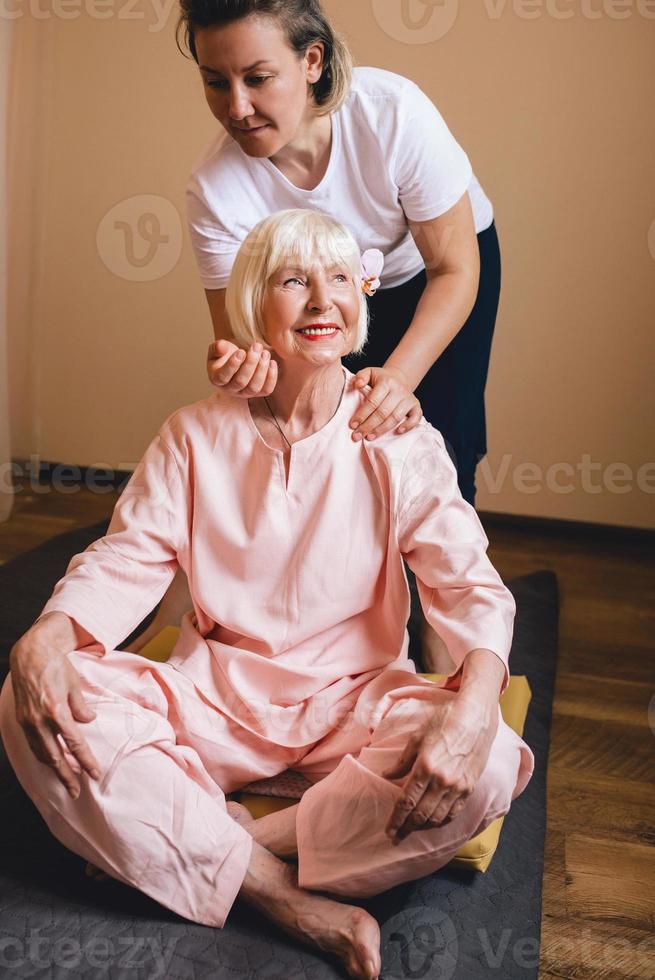 Senior mujer elegante caucásica con cabello gris y phalaenopsis rosa en su cabello sentado en masaje tailandés. anti edad, estilo de vida saludable, viajes, concepto de masaje tailandés foto