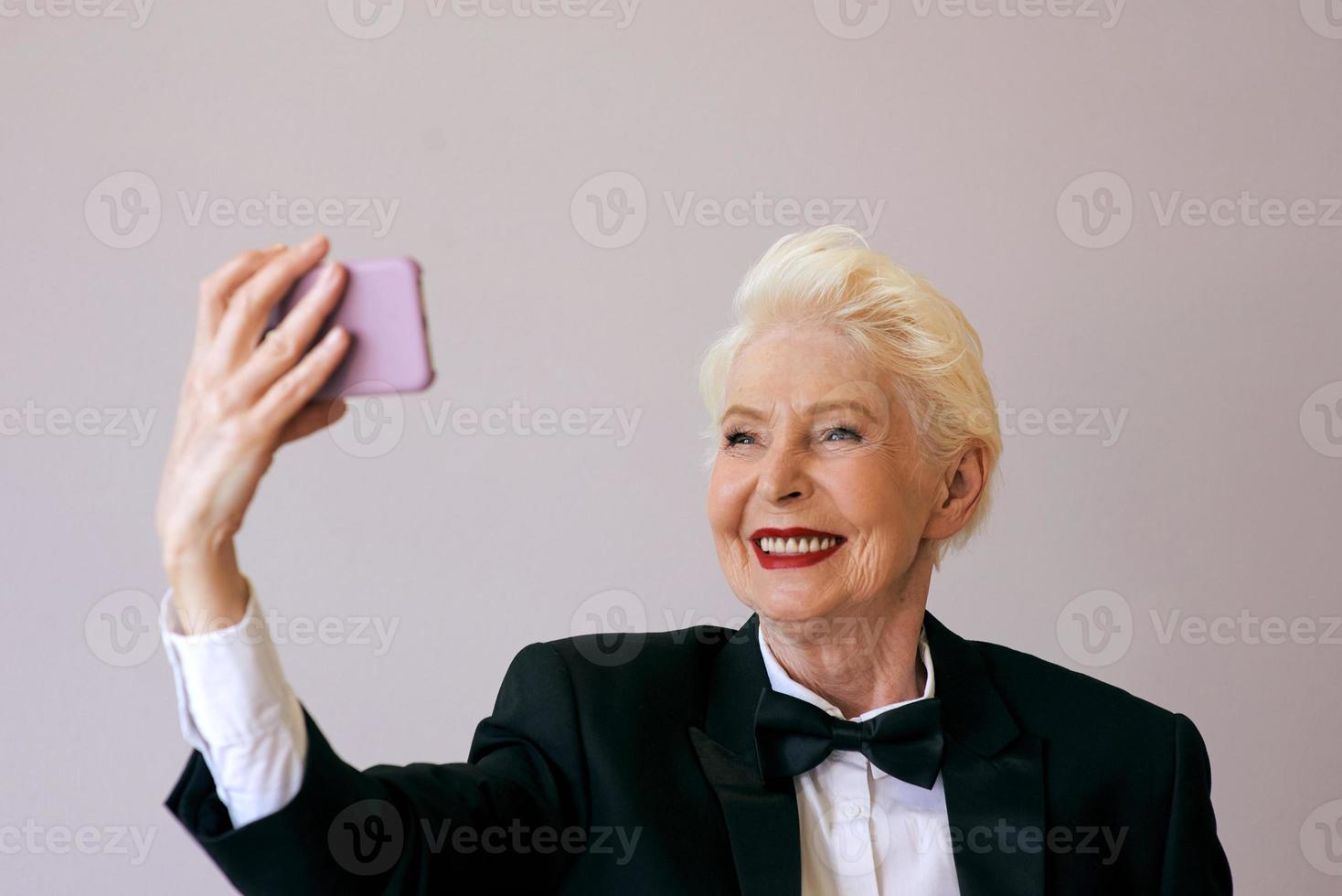 stylish mature senior woman in tuxedo with cellphone video calling or making selfie. Fun, party, style, lifestyle, business, technology, celebration concept photo