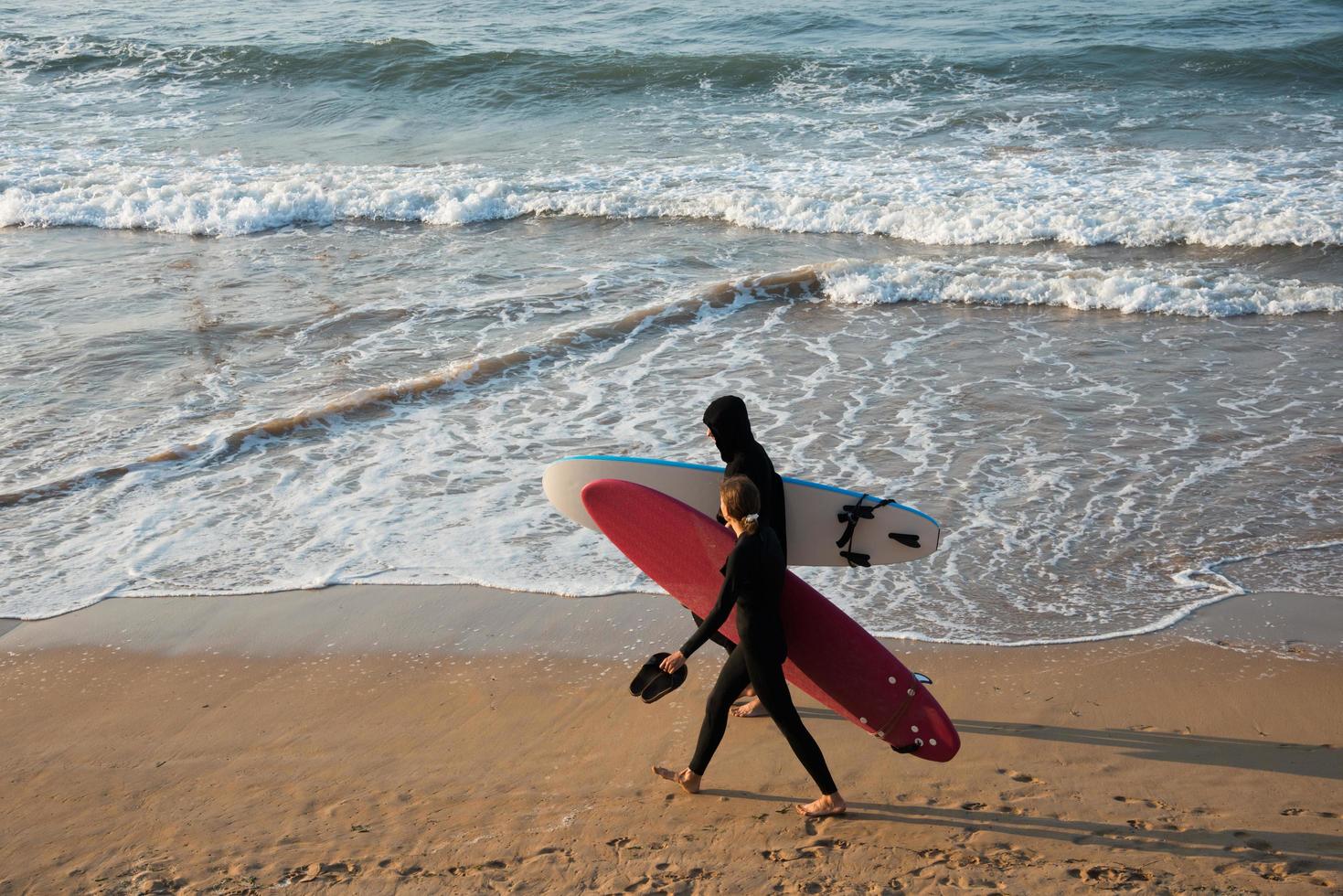 pareja joven caminando por la playa con sus tablas de surf y trajes de neopreno. puesta de sol, playa de san lorenzo, gijón, asturias, españa foto