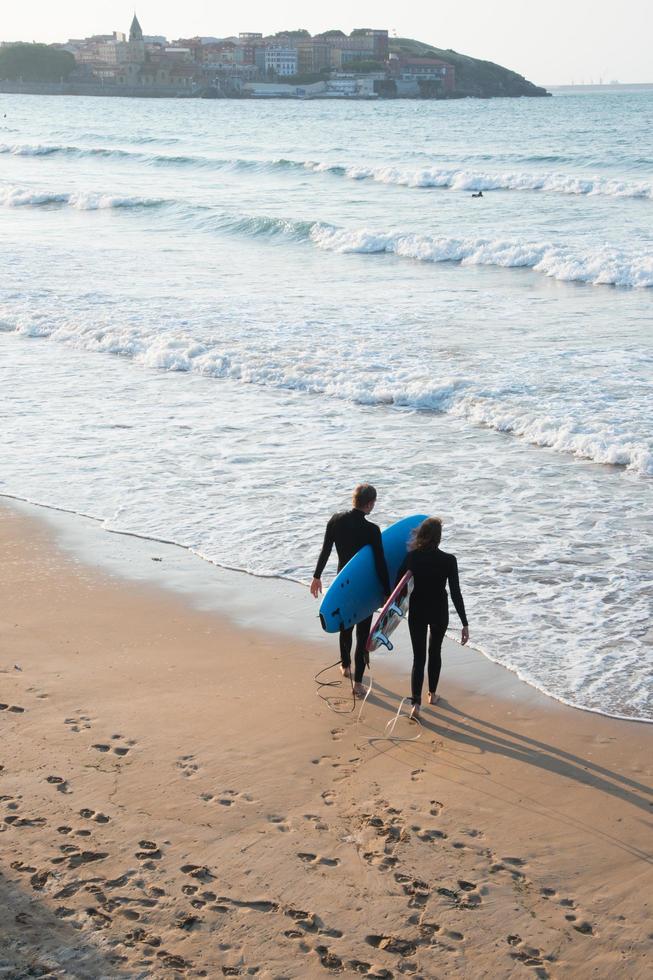 Pareja joven con trajes de neopreno y tablas de surf entran al agua para surfear en la playa de san lorenzo, gijón, españa. visto desde la espalda. foto