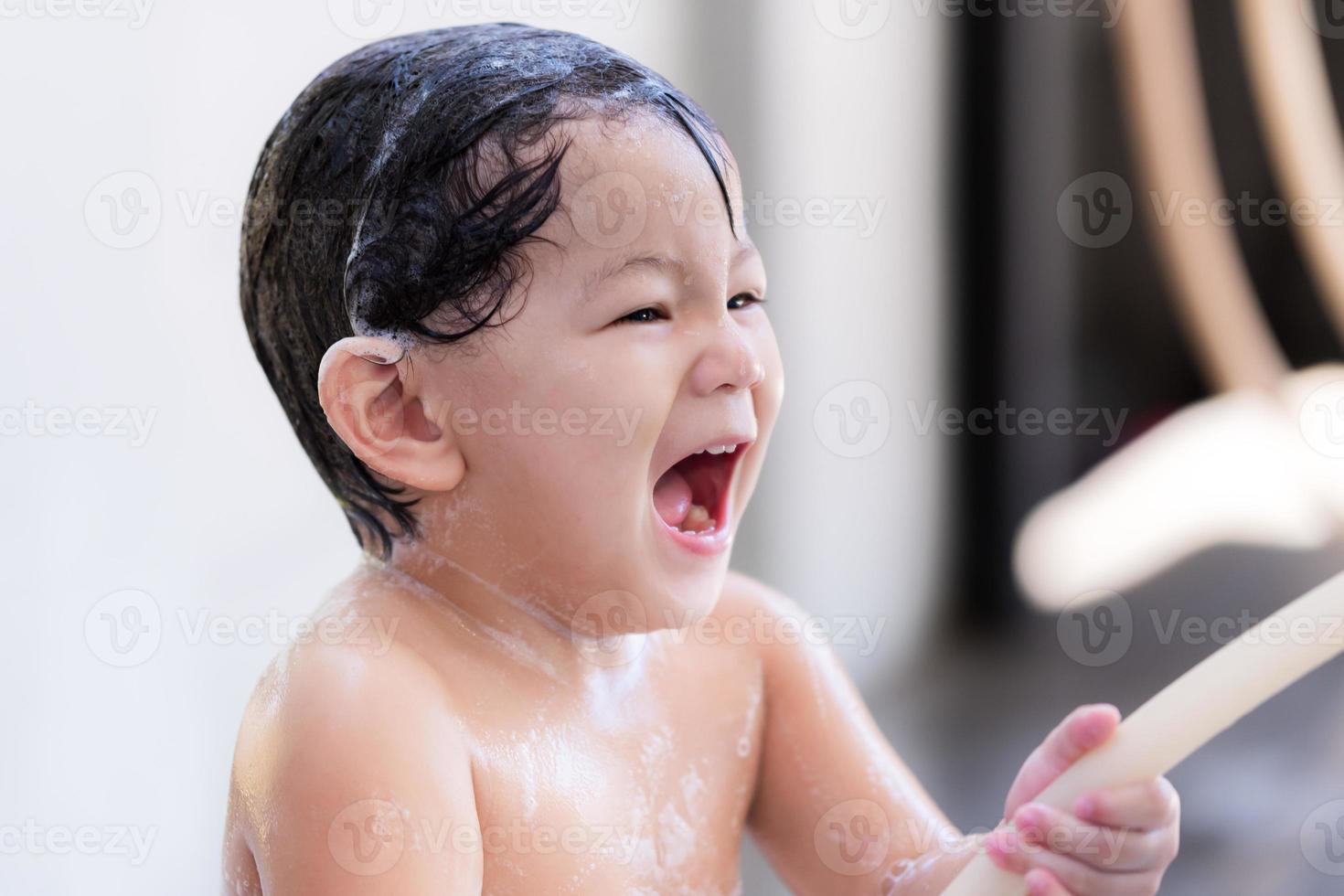 lindo niño preescolar asiático se están riendo. bebé divirtiéndose en bañarse y lavarse el cabello. niño pequeño dulce sonriendo alegre. sus manos sosteniendo una correa de goma para liberar agua limpia. hijo tiene 2 años. foto