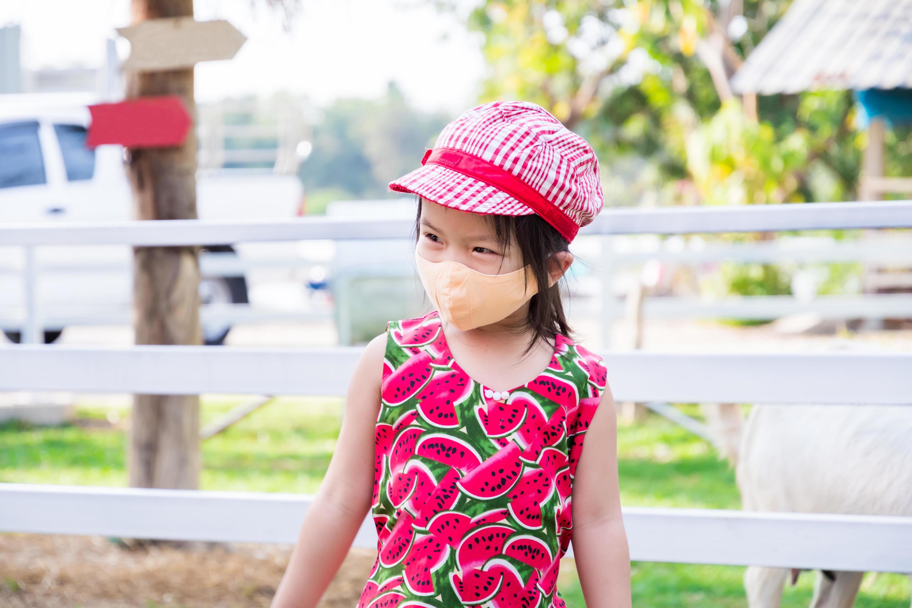 Cute Asian girl wearing orange cloth face mask. Child visit zoo during  coronavirus outbreak. Prevent air pollution PM2.5 dust. Child wear red sun  hood matching dress. Happy kids is 4 years old.