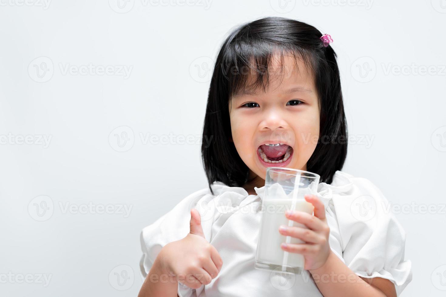 Niña de 4 años está bebiendo leche con vaso. El niño pequeño levantó los pulgares mostrando que estaba bien. fondo blanco aislado. foto