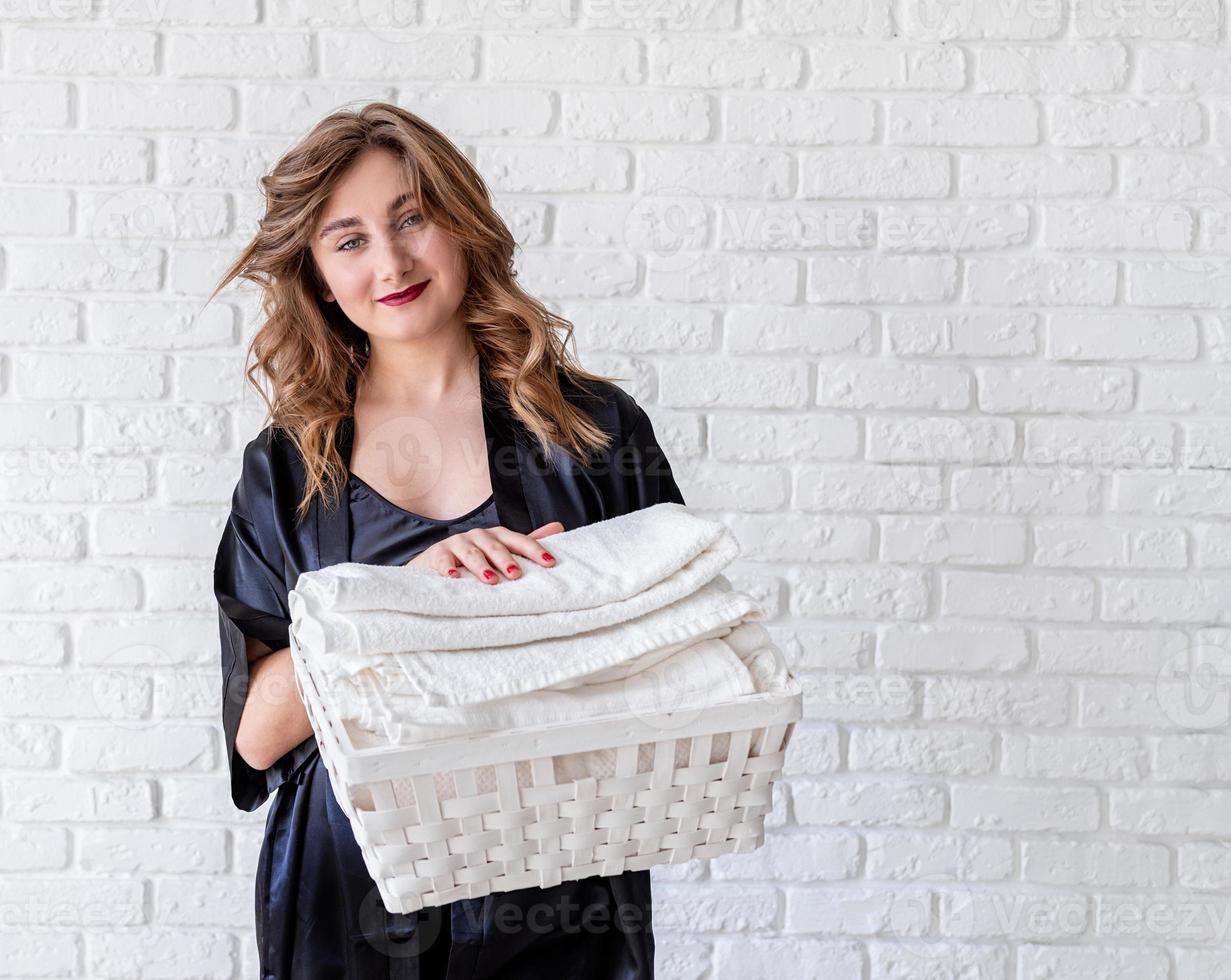 Mujer sonriente sosteniendo una canasta blanca con toallas sobre fondo de ladrillos blancos con espacio de copia foto