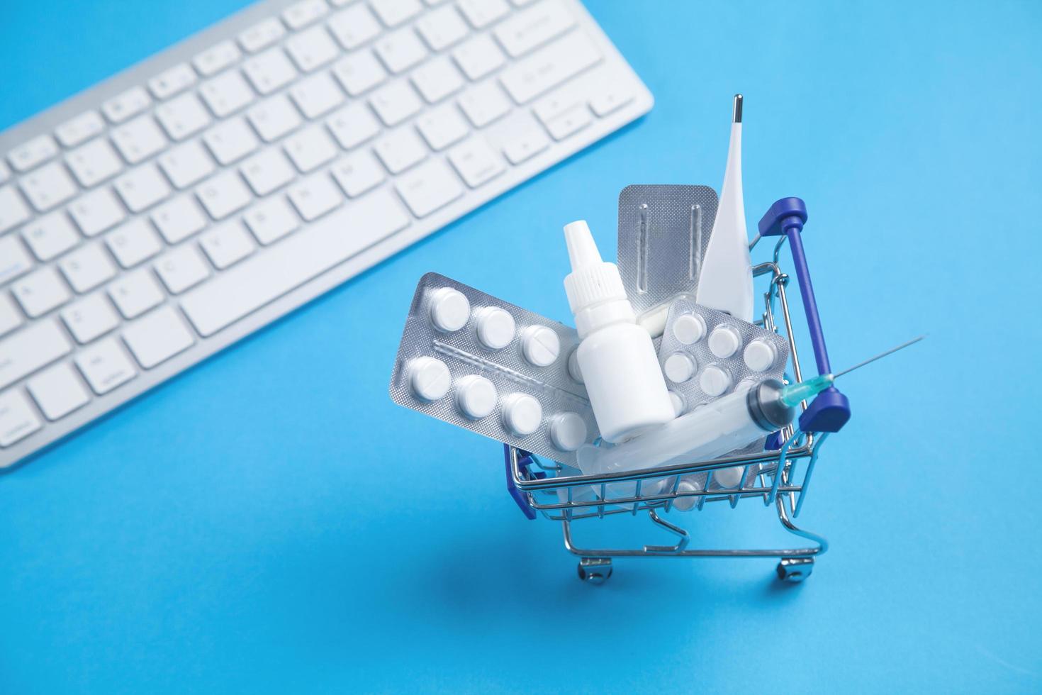 Shopping cart with pills, medical bottle, thermometer, syringe and keyboard on a blue background. photo