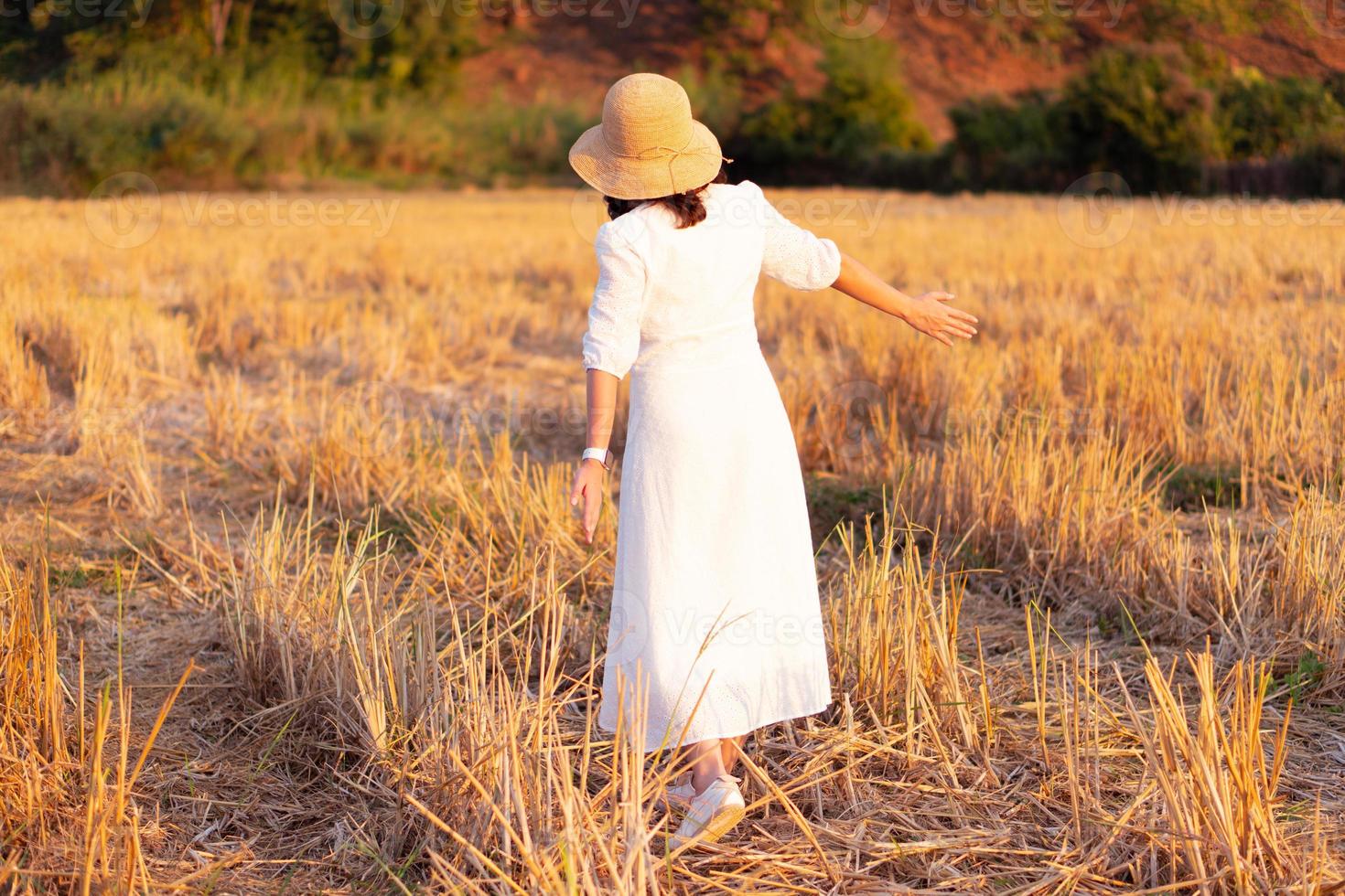 portrait of young woman wearing straw hat and white dress walking in dry rice paddy field at sunset photo