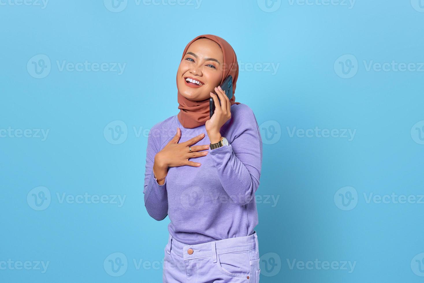 Smiling young Asian woman talking on mobile phone and put hands chest on blue background photo