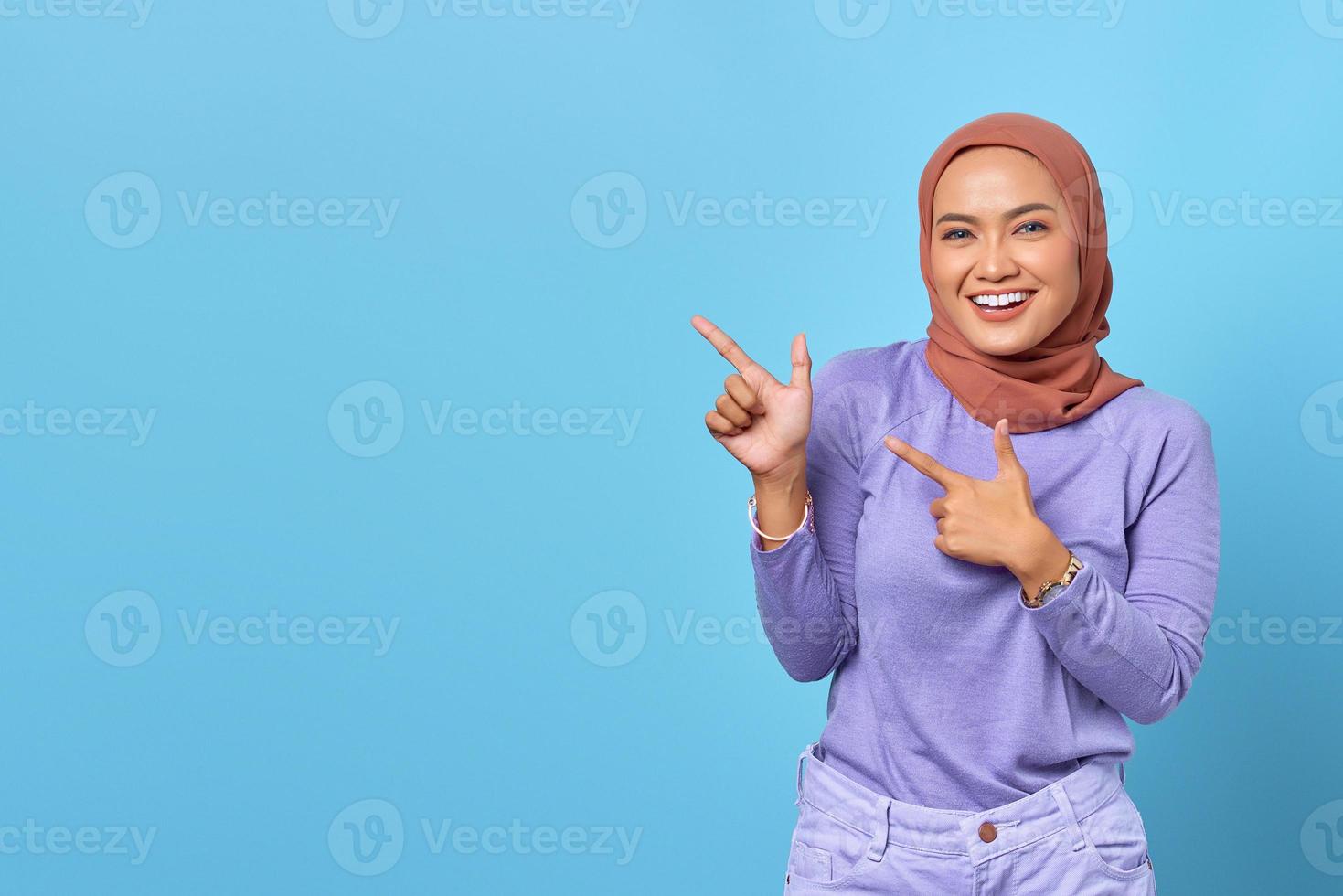 Portrait of smiling young Asian woman pointing finger at copy space on blue background photo