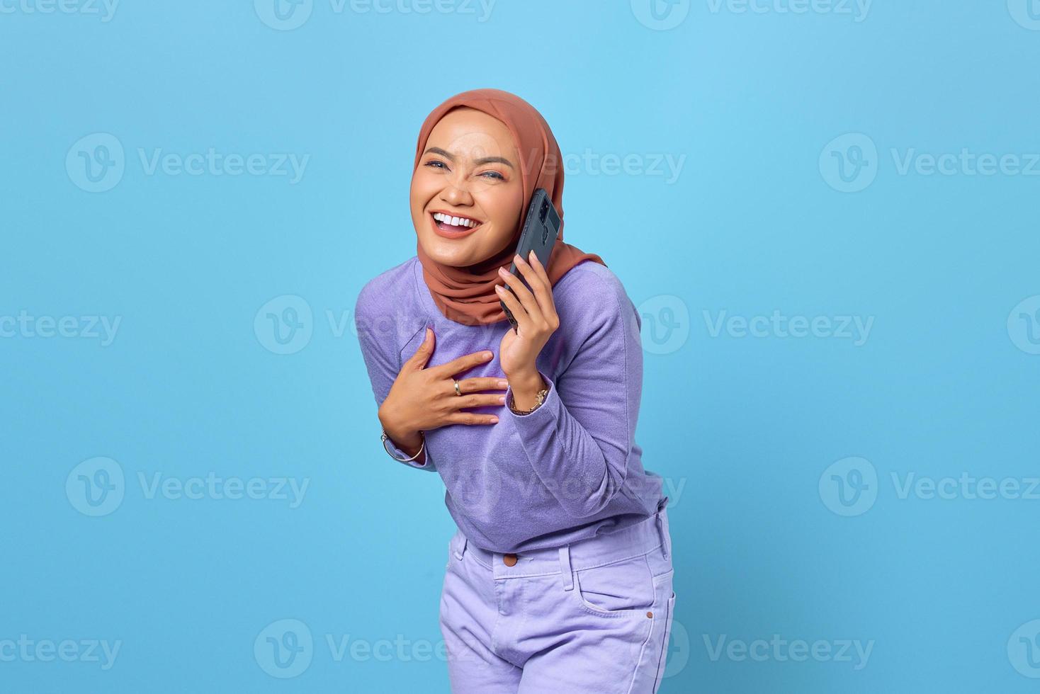 Sonriente joven mujer asiática hablando por teléfono móvil y poner las manos en el pecho sobre fondo azul. foto