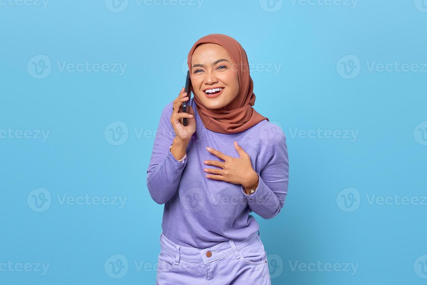 Sonriente joven mujer asiática hablando por teléfono móvil y poner las manos en el pecho sobre fondo azul. foto