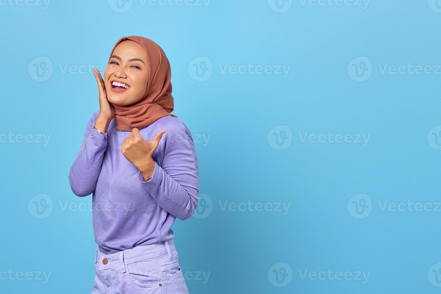 Portrait of smiling young Asian woman pointing a thumb at copy space on blue background photo