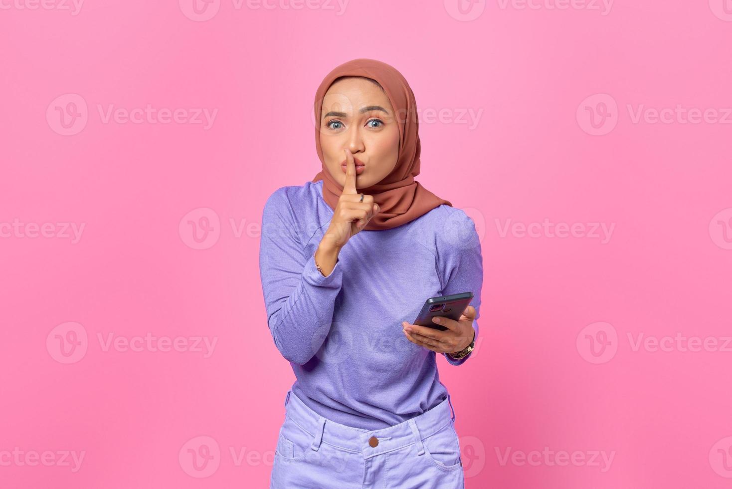 Beautiful young Asian woman holding mobile phone and makes silence gesture on pink background photo