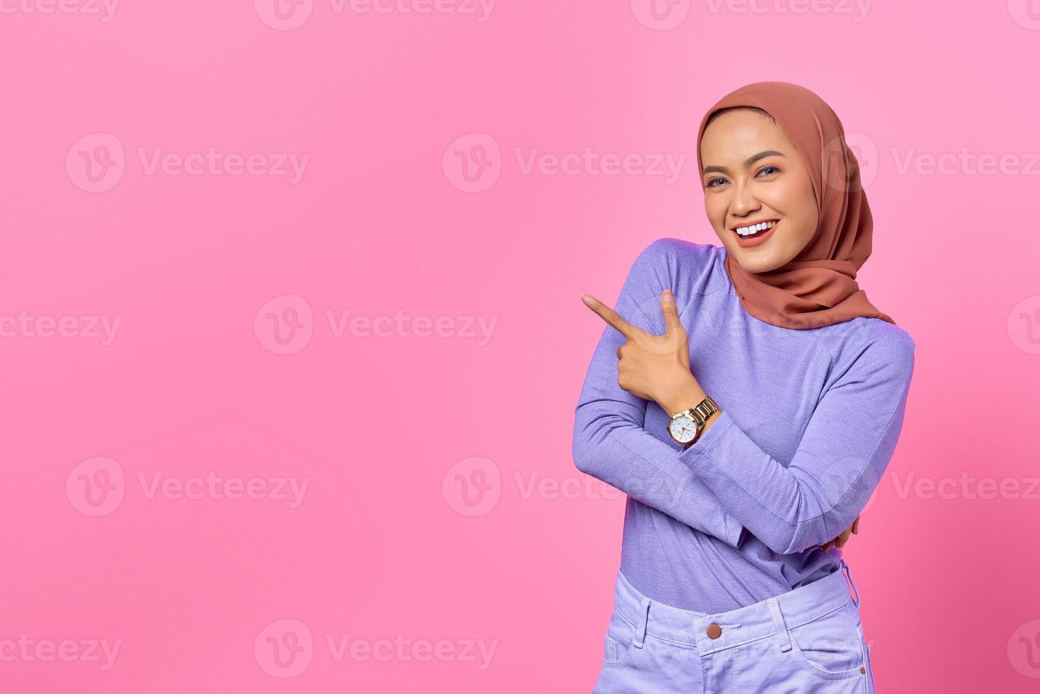 Portrait of smiling young Asian woman pointing finger at copy space on pink background photo