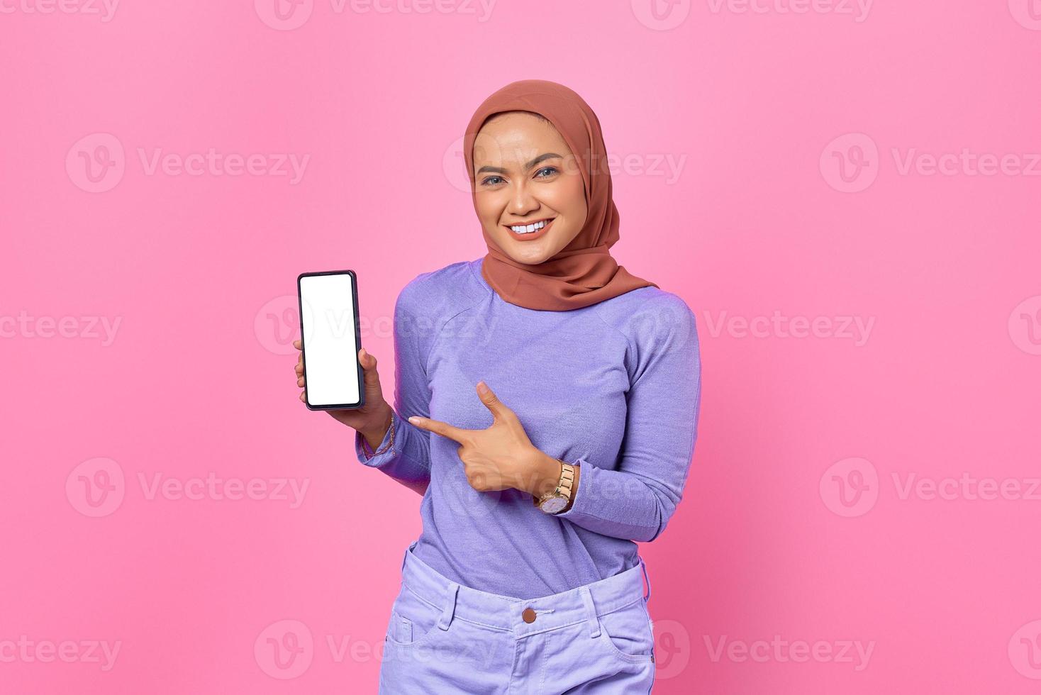 Portrait of smiling young Asian woman pointing finger on a mobile phone screen on pink background photo