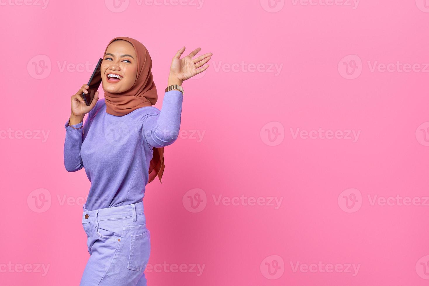 Sonriente joven mujer asiática hablando por teléfono móvil mientras agita el gesto de la mano sobre fondo rosa foto