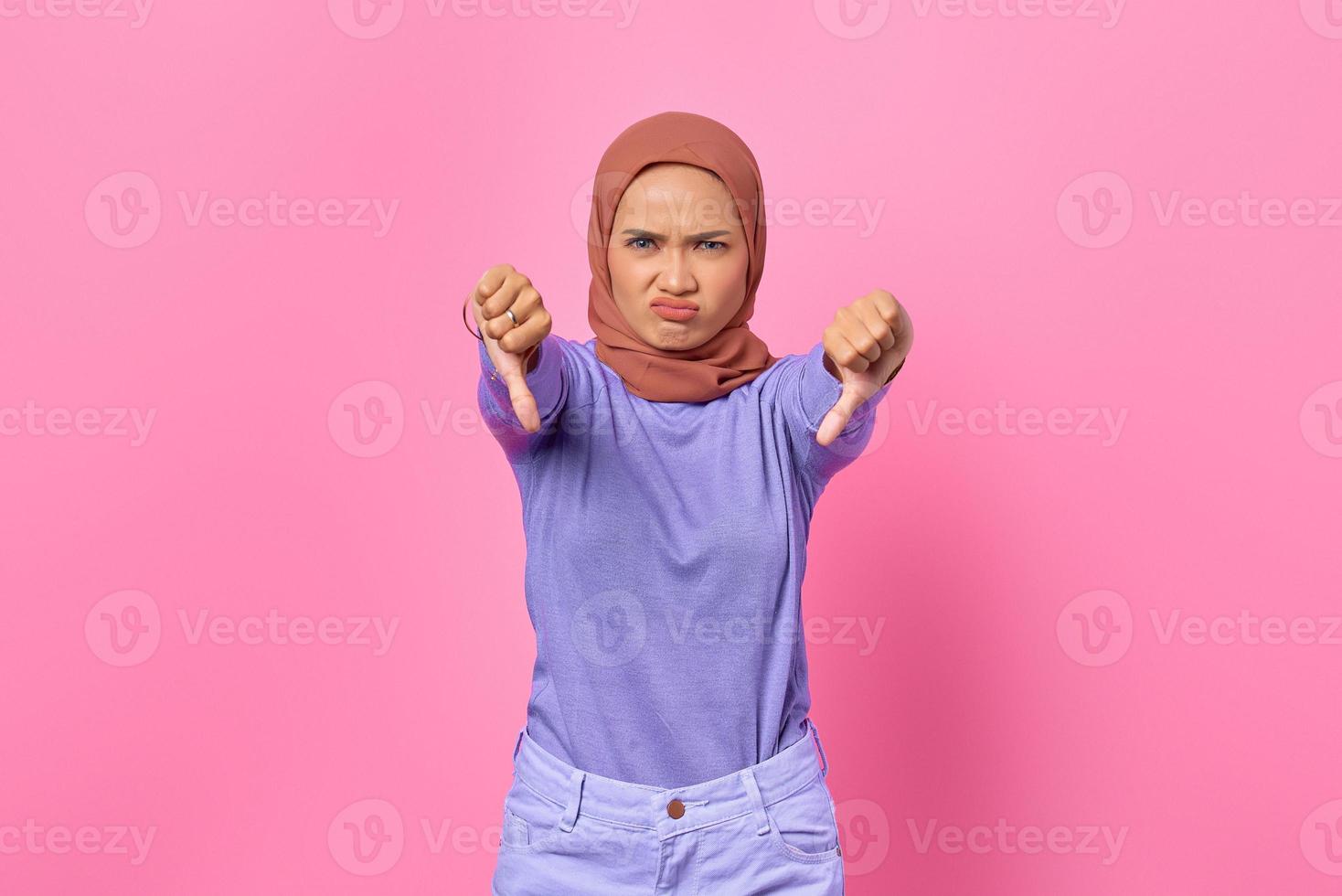 enojado, joven, mujer asiática, actuación, rechazo, con, pulgares abajo, gesto, en, fondo rosa foto