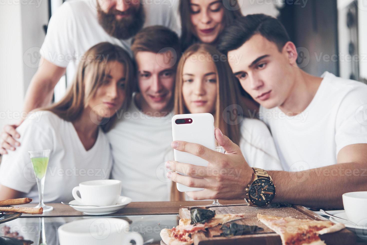 A group of people make a selfie photo in a cafe. The best friends gathered together at a dinner table eating pizza and singing various drinks