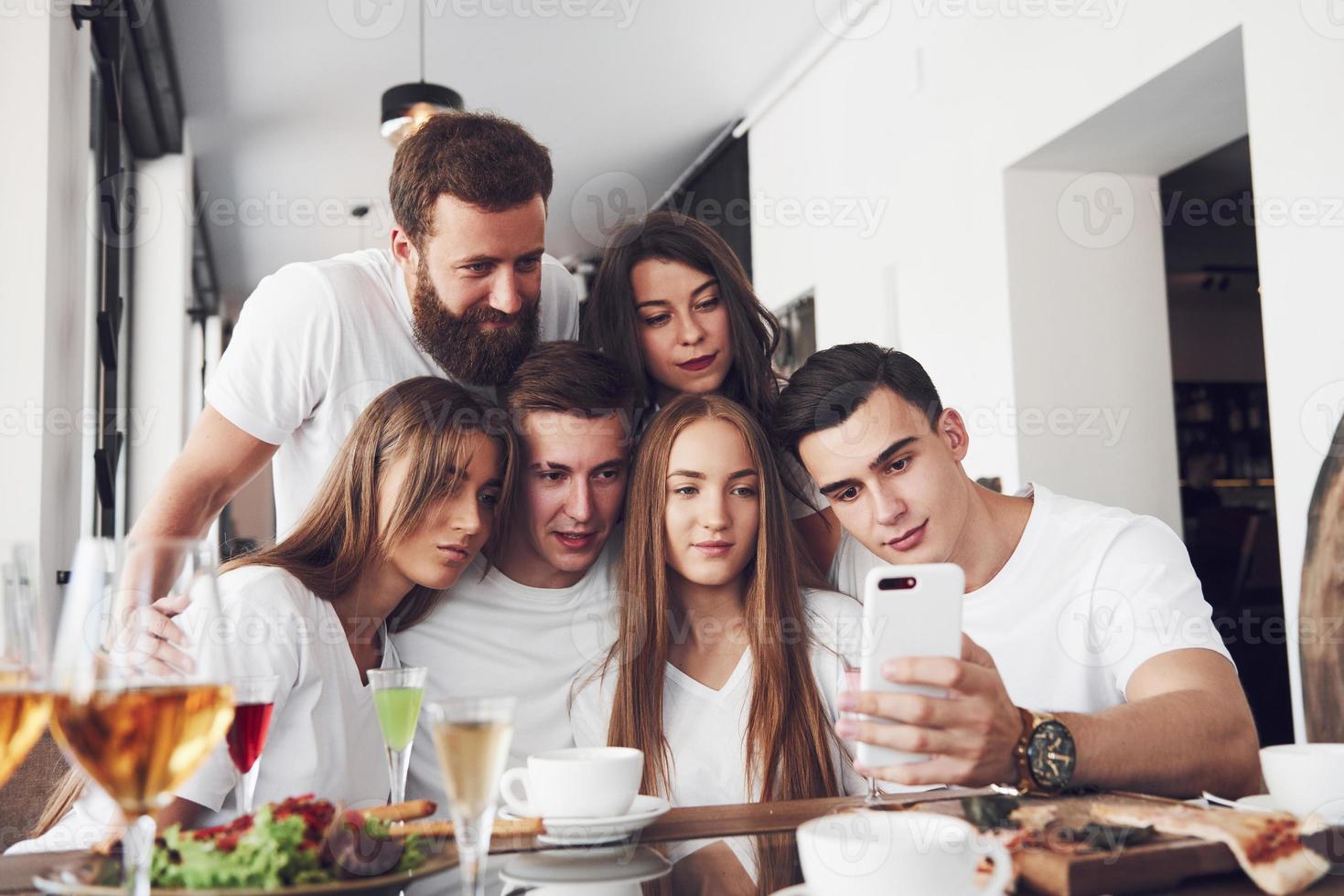 A group of people make a selfie photo in a cafe. The best friends gathered together at a dinner table eating pizza and singing various drinks