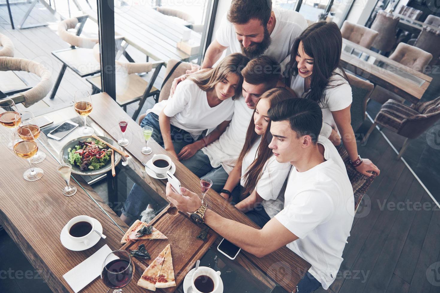 A group of people make a selfie photo in a cafe. The best friends gathered together at a dinner table eating pizza and singing various drinks