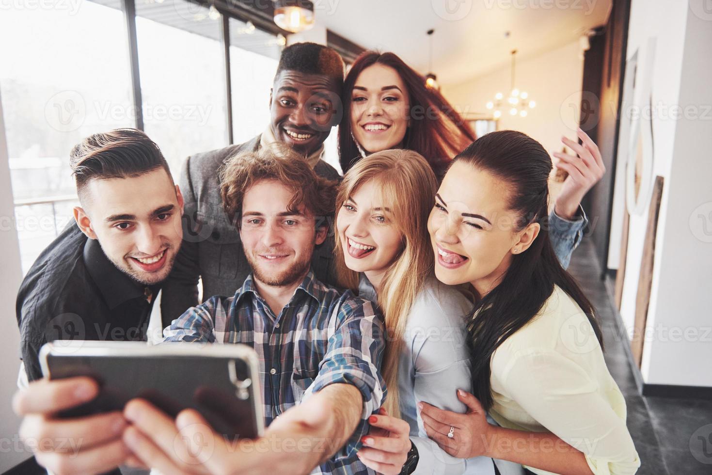 Friends having fun at restaurant. Three boys and three girls making selfie and laughing. On foreground boy holding smart phone. All wear casual clothes photo