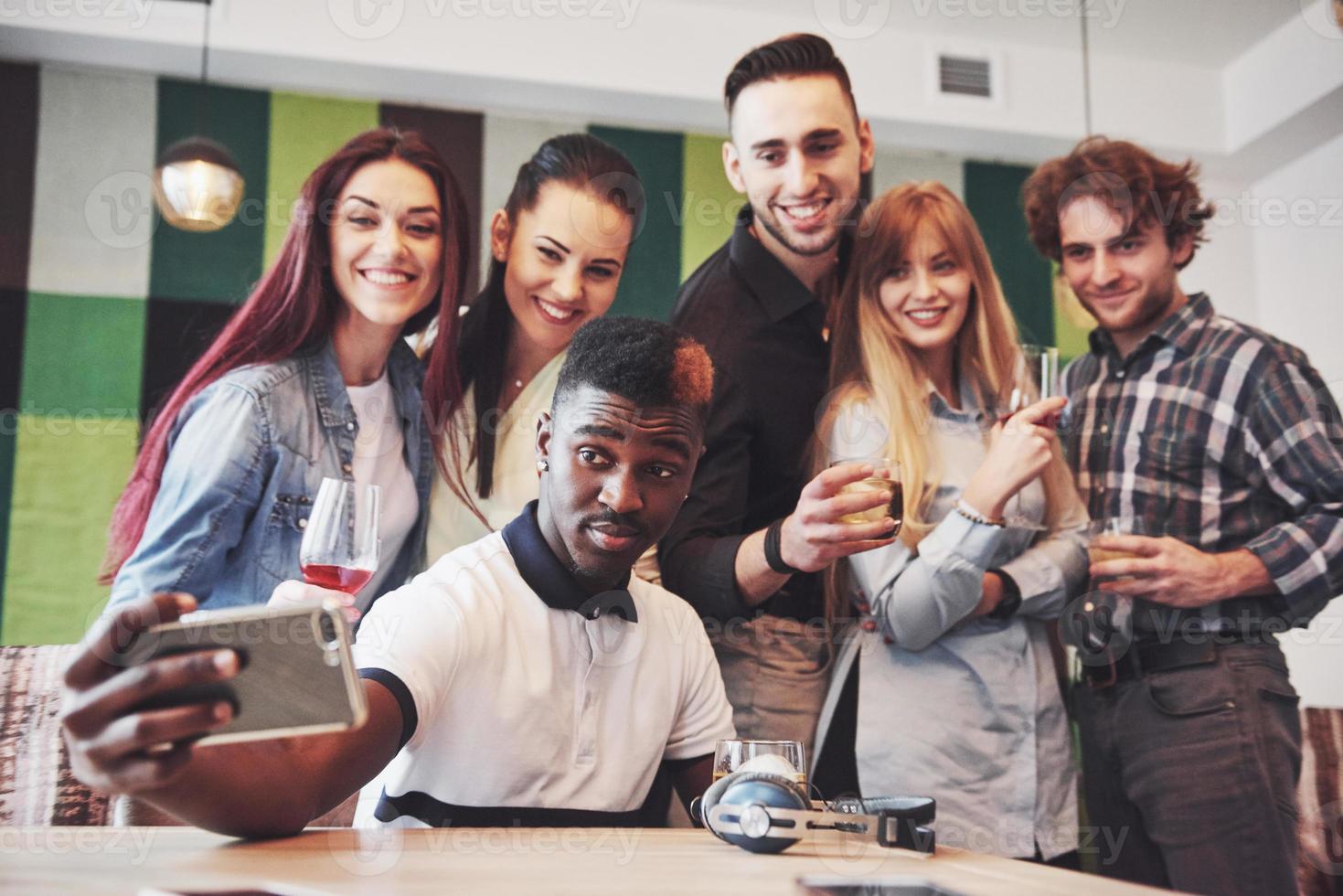 Friends having fun at restaurant. Three boys and three girls making selfie and laughing. On foreground boy holding smart phone. All wear casual clothes photo