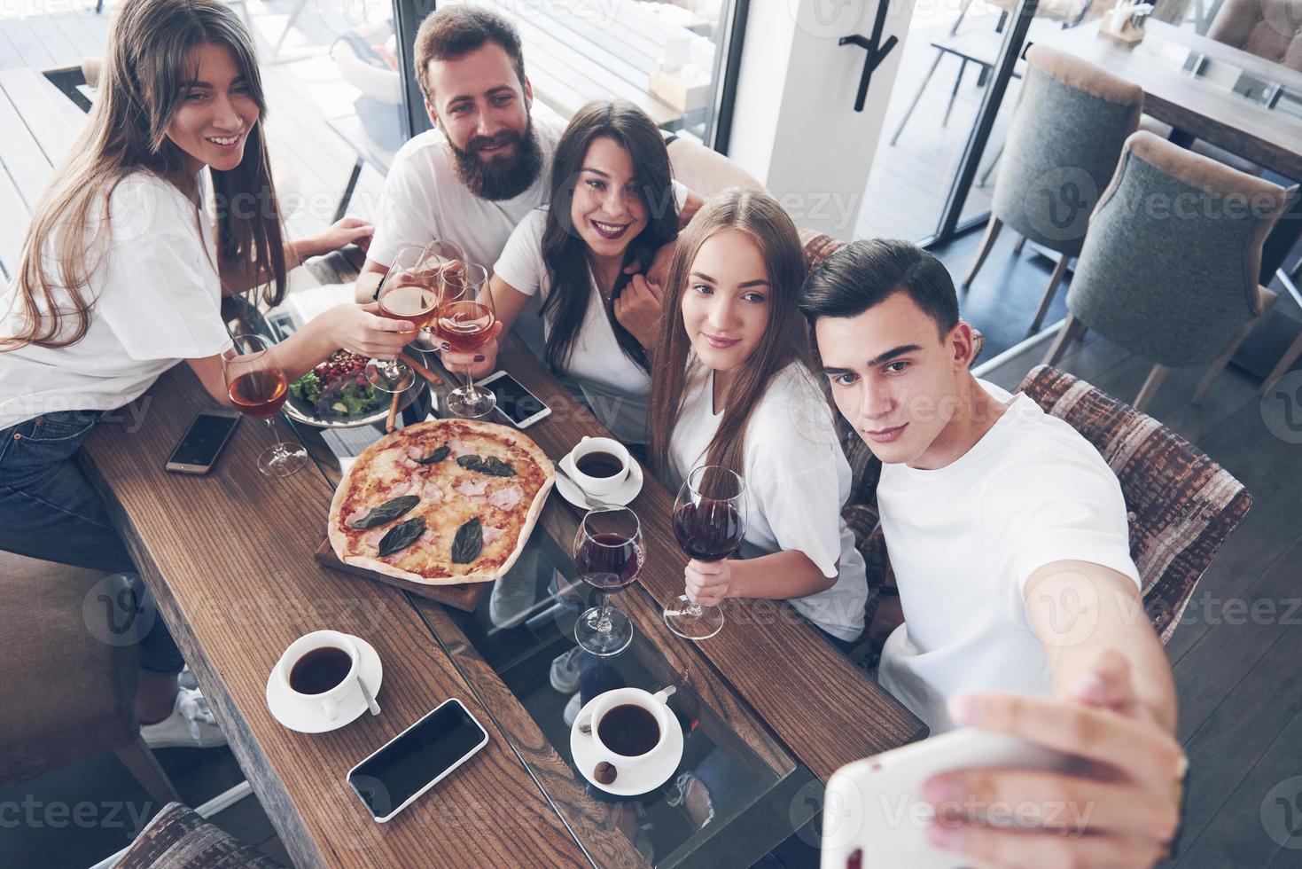 A group of people make a selfie photo in a cafe. The best friends gathered together at a dinner table eating pizza and singing various drinks