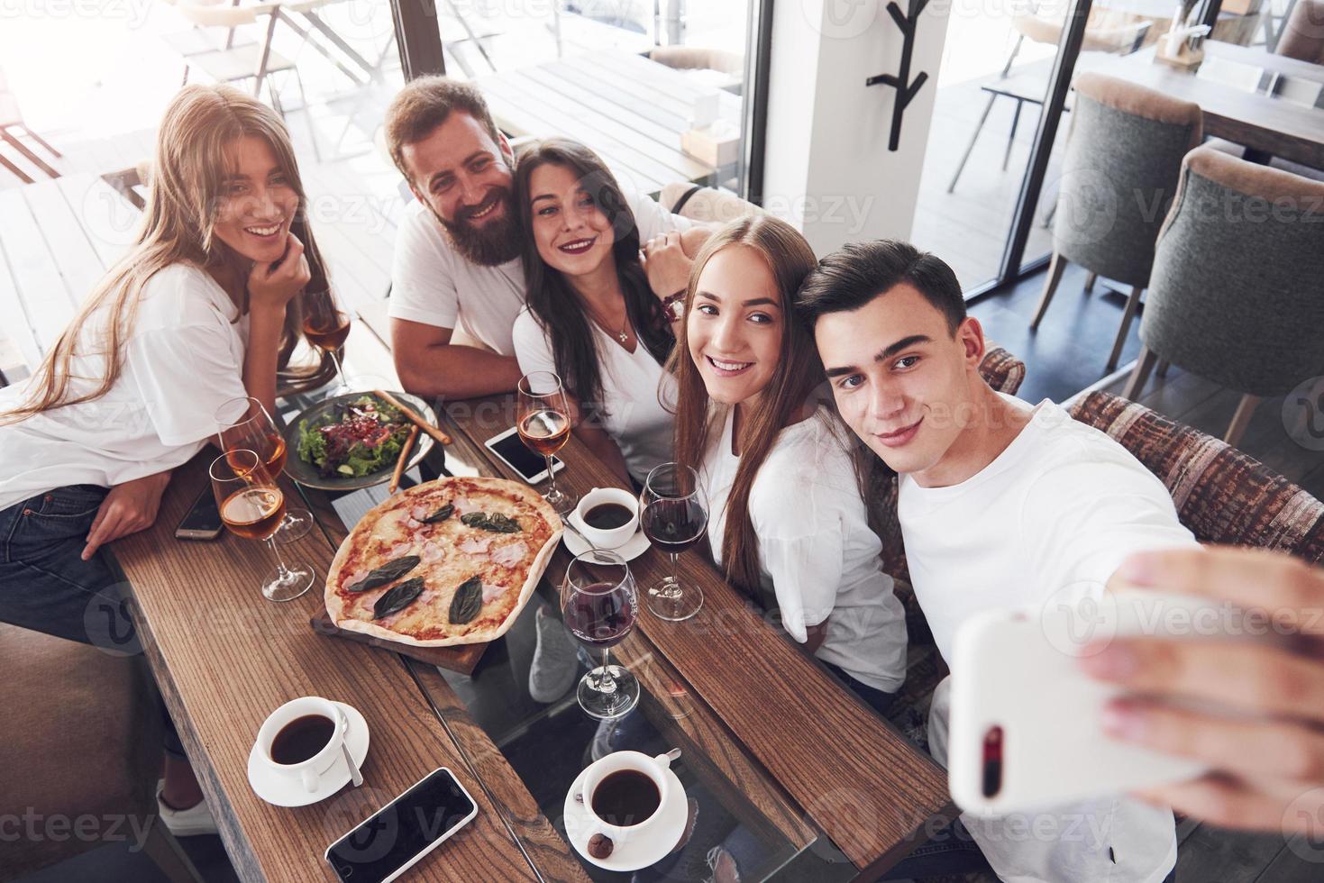 A group of people make a selfie photo in a cafe. The best friends gathered together at a dinner table eating pizza and singing various drinks