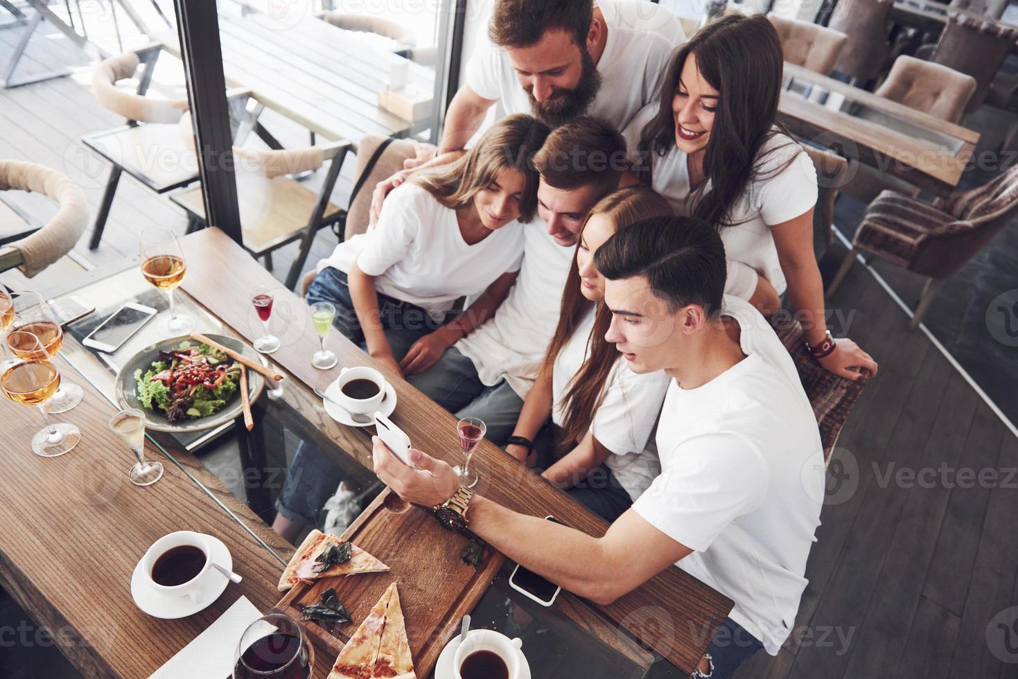 A group of people make a selfie photo in a cafe. The best friends gathered together at a dinner table eating pizza and singing various drinks