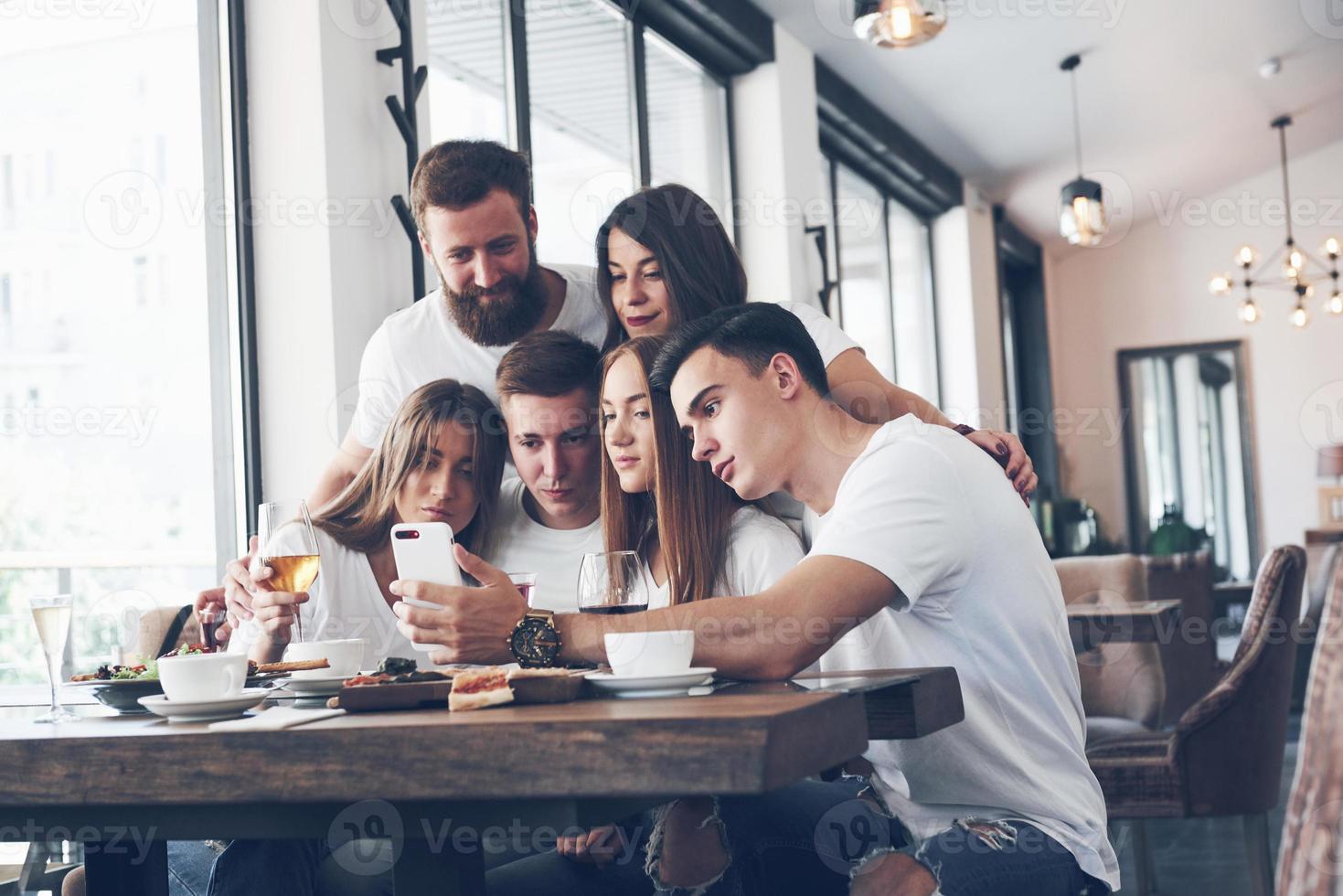 A group of people make a selfie photo in a cafe. The best friends gathered together at a dinner table eating pizza and singing various drinks