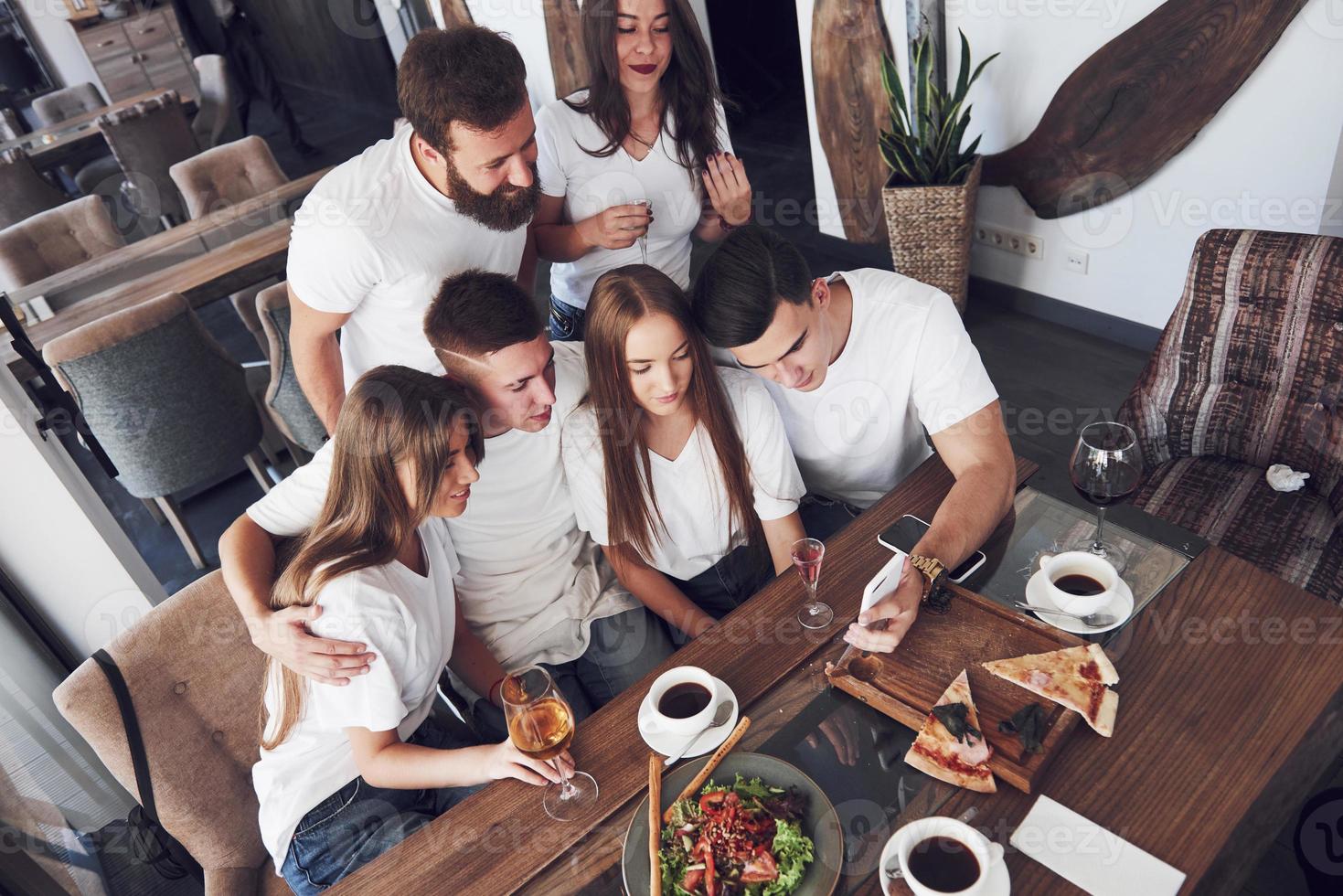 A group of people make a selfie photo in a cafe. The best friends gathered together at a dinner table eating pizza and singing various drinks