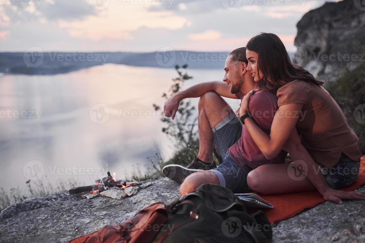 pareja abrazándose con mochila sentado cerca del fuego en la cima de la montaña disfrutando de la vista de la costa, un río o un lago. viajando a lo largo de las montañas y la costa, la libertad y el concepto de estilo de vida activo foto