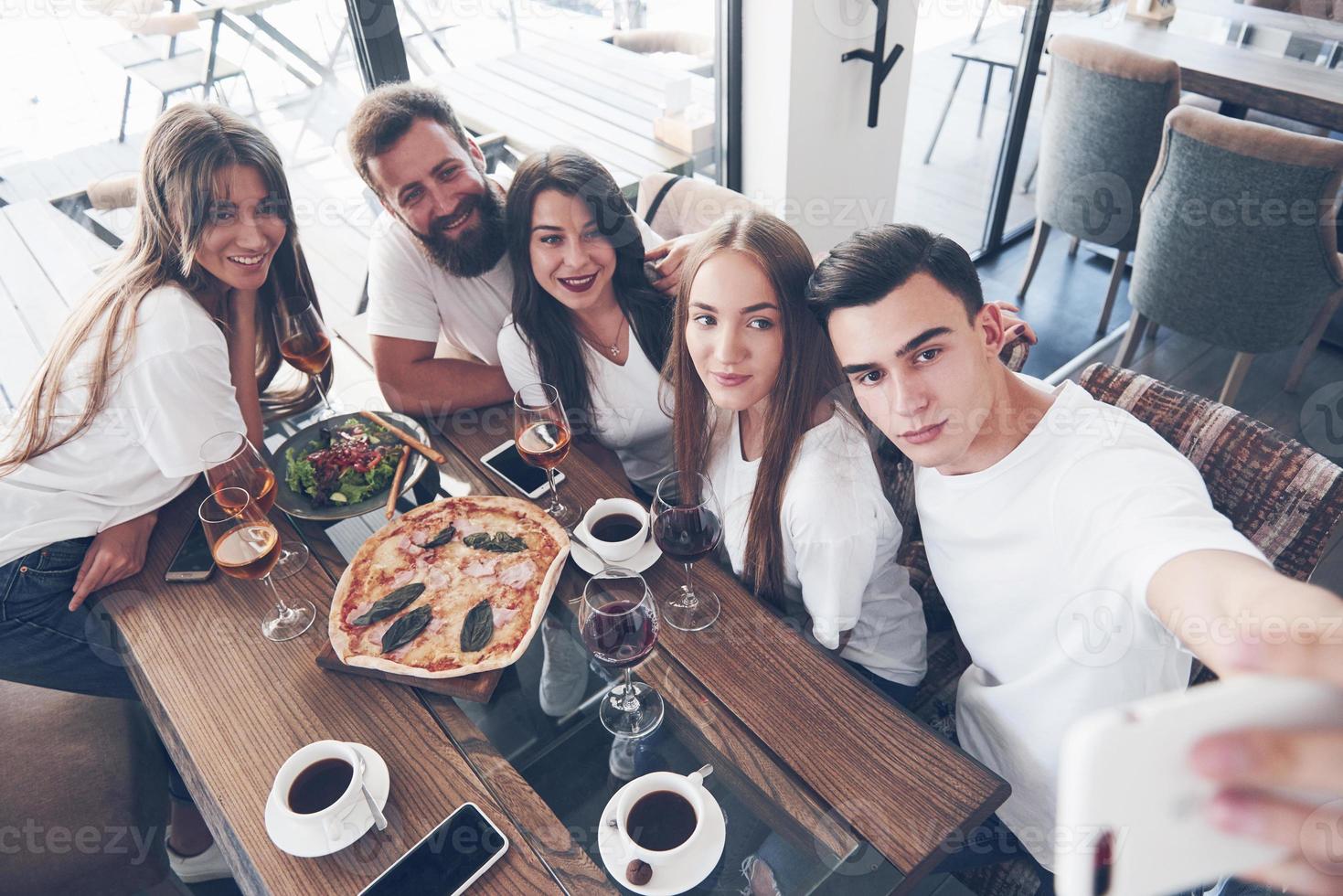 A group of people make a selfie photo in a cafe. The best friends gathered together at a dinner table eating pizza and singing various drinks