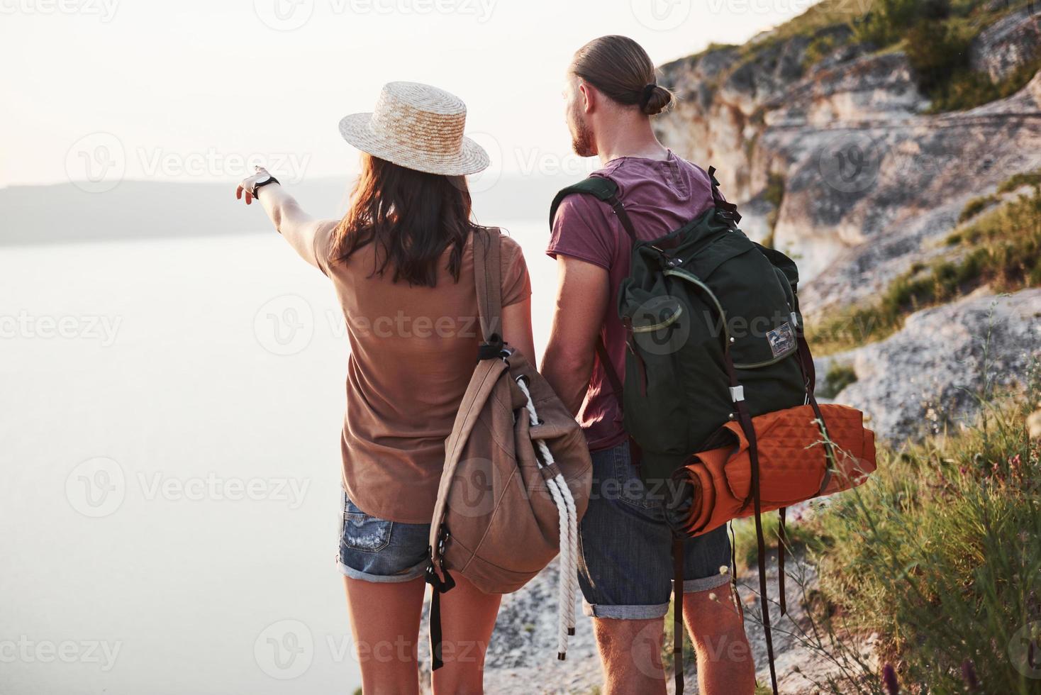 Two tourist male and woman with backpacks stand to the top of the crag and enjoying sunrise. Traveling mountains and coast, freedom and active lifestyle concept photo