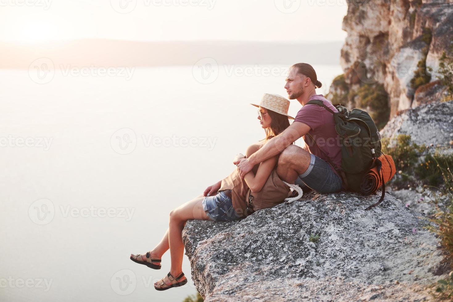 pareja abrazándose con mochila sentado en la cima de la montaña de roca disfrutando de la vista de la costa, un río o un lago. viajando a lo largo de las montañas y la costa, la libertad y el concepto de estilo de vida activo foto