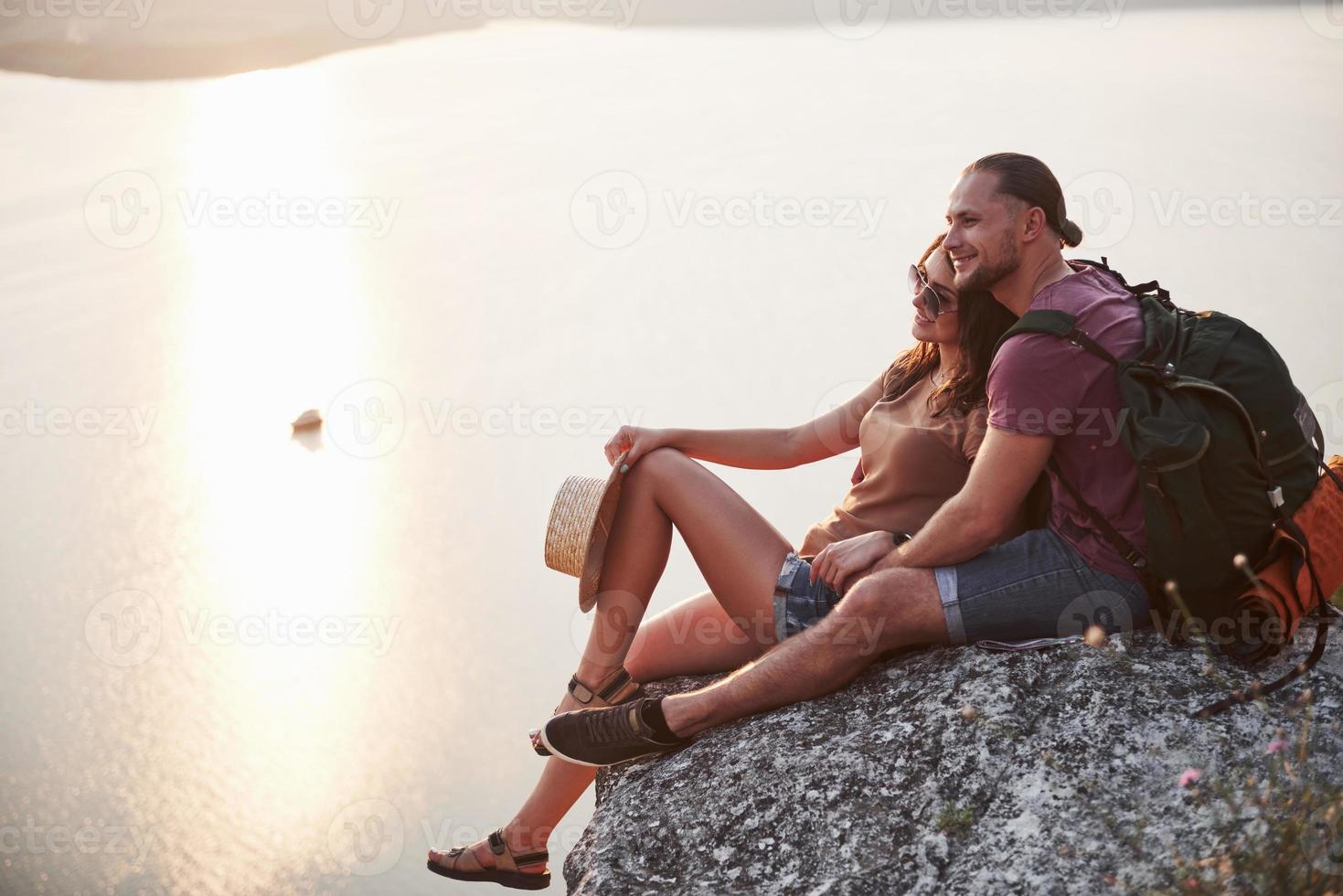 Hugging couple with backpack sitting on top of rock mountain enjoying view coast a river or lake. Traveling along mountains and coast, freedom and active lifestyle concept photo