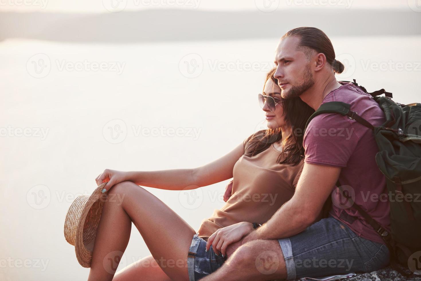 Hugging couple with backpack sitting on top of rock mountain enjoying view coast a river or lake. Traveling along mountains and coast, freedom and active lifestyle concept photo