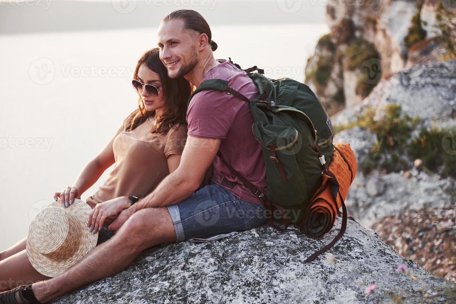 pareja abrazándose con mochila sentado en la cima de la montaña de roca disfrutando de la vista de la costa, un río o un lago. viajando a lo largo de las montañas y la costa, la libertad y el concepto de estilo de vida activo foto