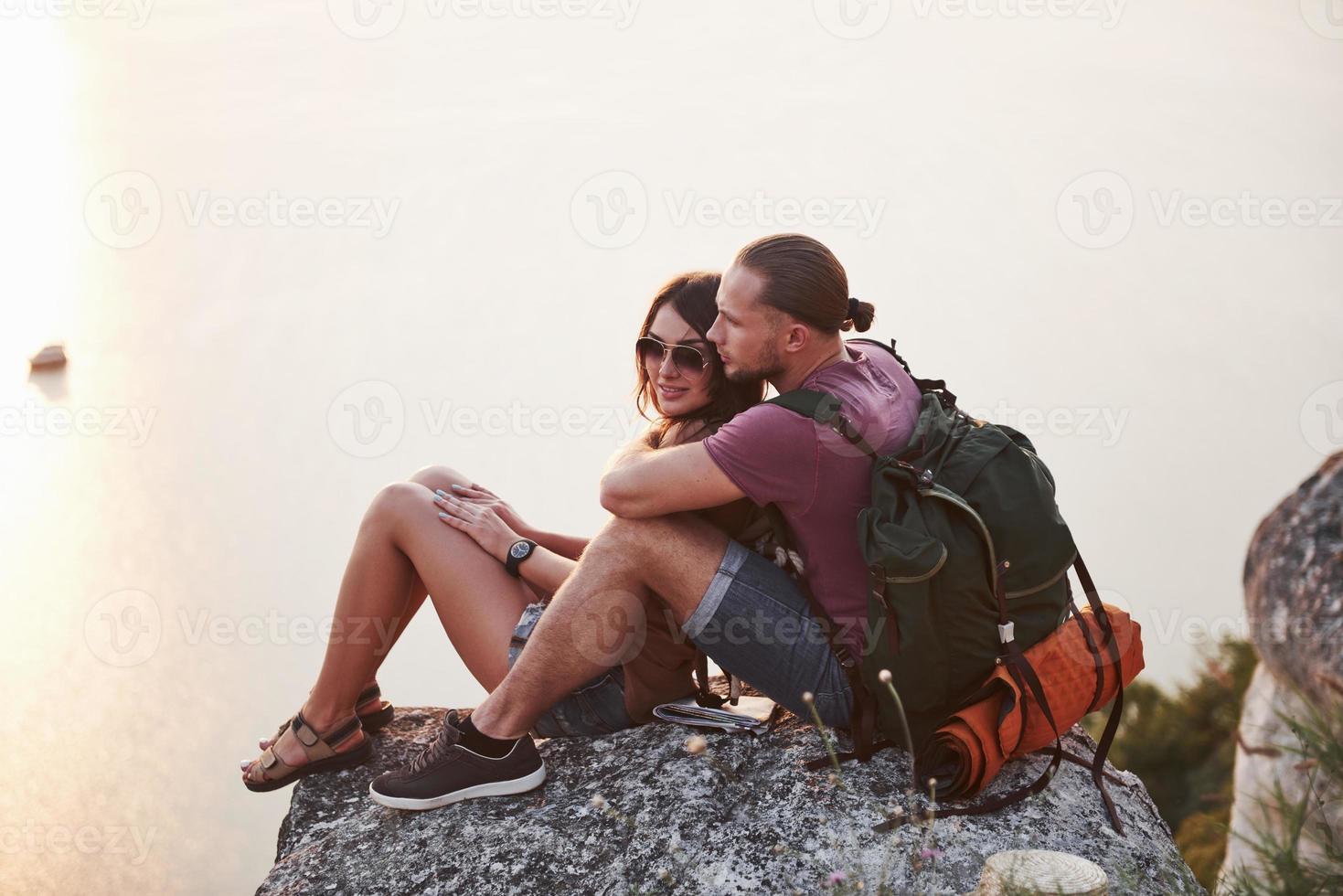 Hugging couple with backpack sitting on top of rock mountain enjoying view coast a river or lake. Traveling along mountains and coast, freedom and active lifestyle concept photo