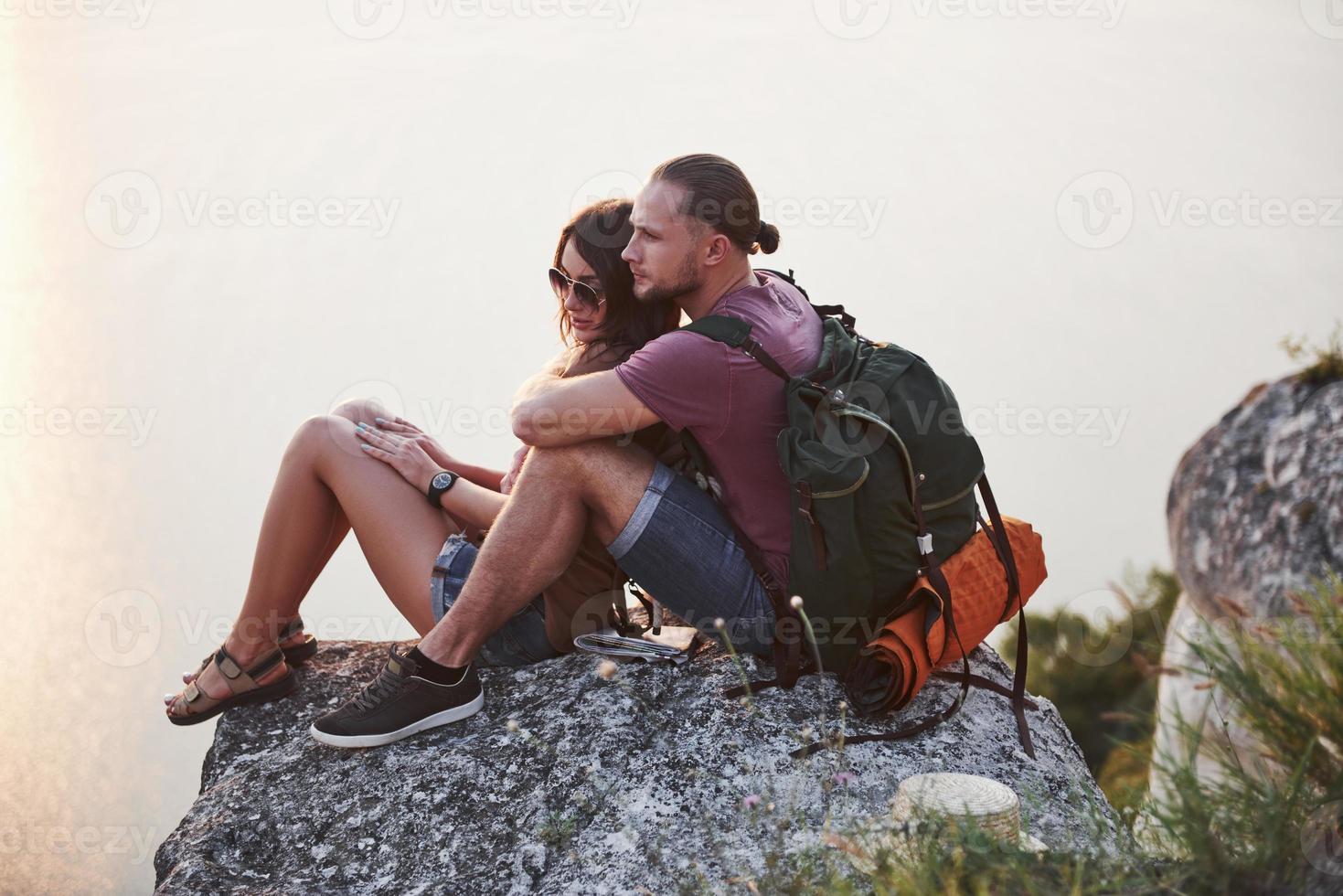 Hugging couple with backpack sitting on top of rock mountain enjoying view coast a river or lake. Traveling along mountains and coast, freedom and active lifestyle concept photo