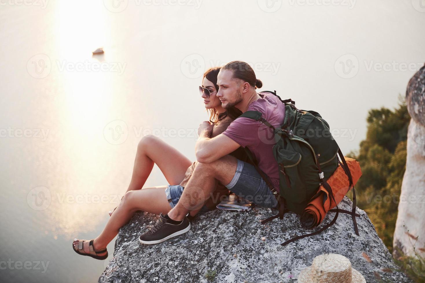 Hugging couple with backpack sitting on top of rock mountain enjoying view coast a river or lake. Traveling along mountains and coast, freedom and active lifestyle concept photo