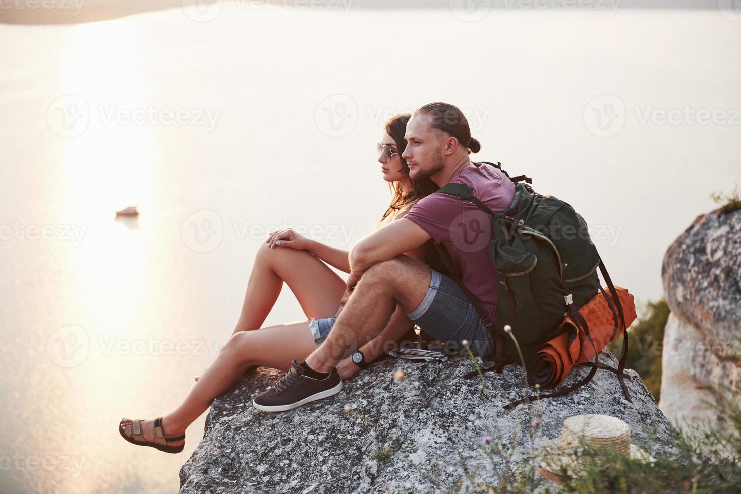 Hugging couple with backpack sitting on top of rock mountain enjoying view coast a river or lake. Traveling along mountains and coast, freedom and active lifestyle concept photo