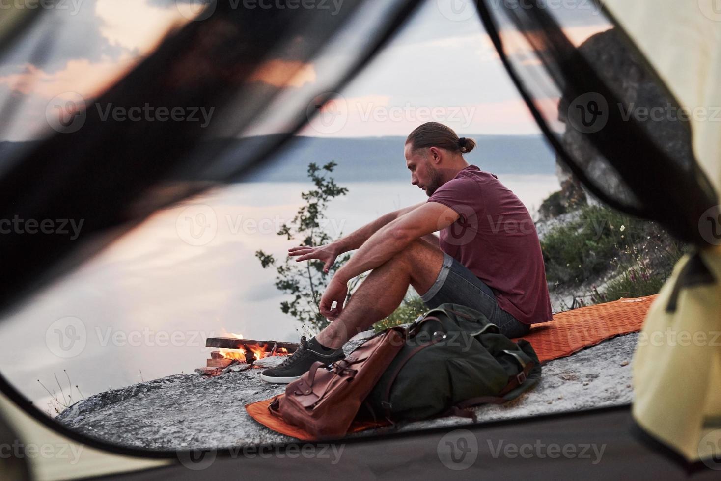 View from tent of traveller with backpack sitting on top of mountain enjoying view coast a river or lake. Traveling along mountains and coast, freedom and active lifestyle concept photo