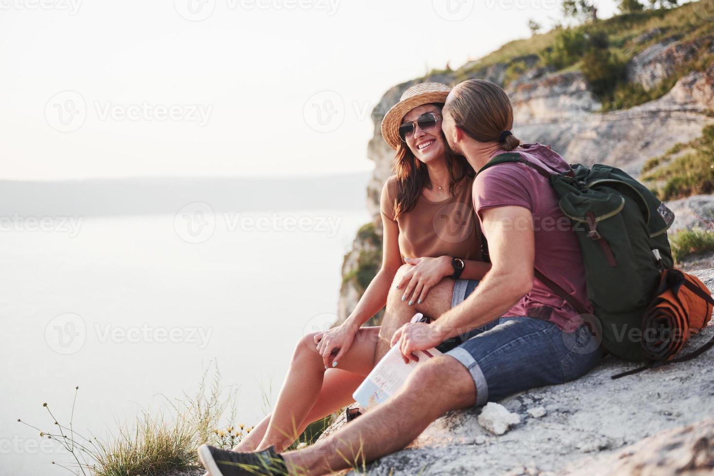 Hugging couple with backpack sitting on top of rock mountain enjoying view coast a river or lake. Traveling along mountains and coast, freedom and active lifestyle concept photo