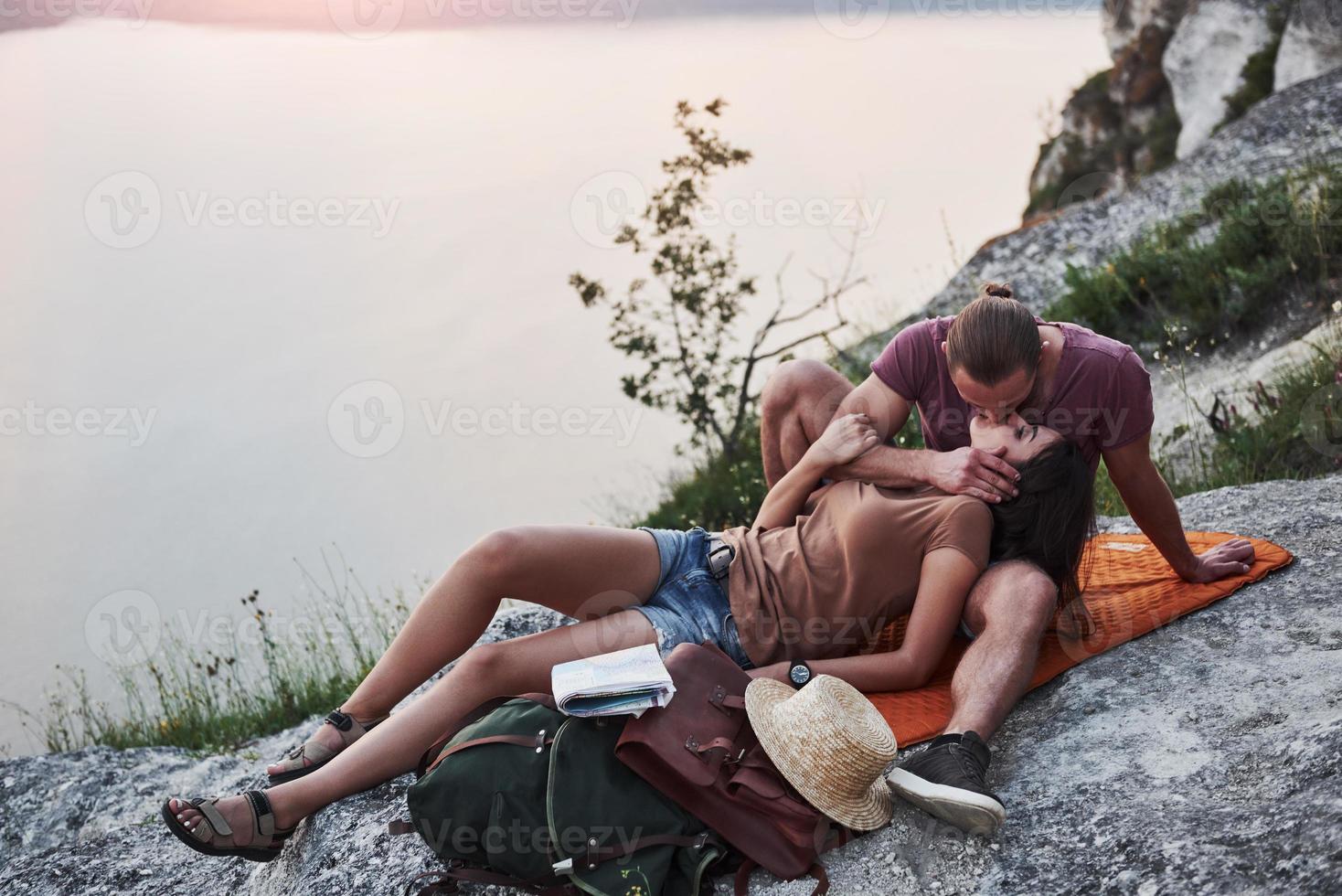 Hugging couple with backpack sitting on top of rock mountain enjoying view coast a river or lake. Traveling along mountains and coast, freedom and active lifestyle concept photo